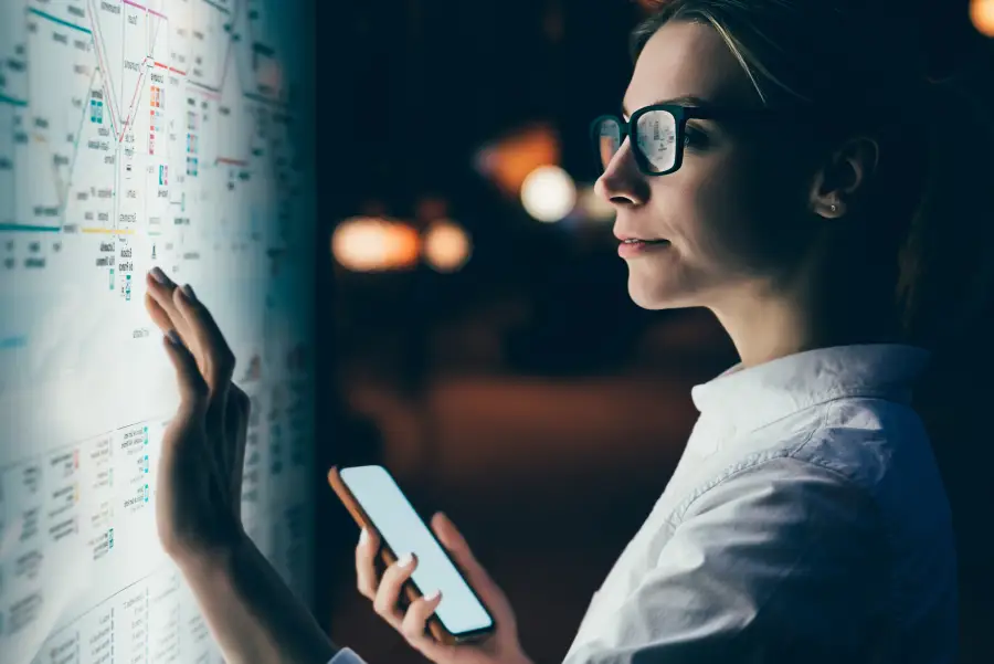 Woman in glasses holding a smartphone while pointing at a lit transit map display in a dark environment.