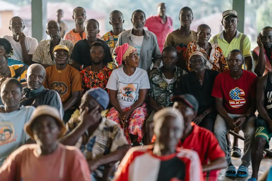 A large group of men and women of various ages seated and standing together at an outdoor community meeting.
