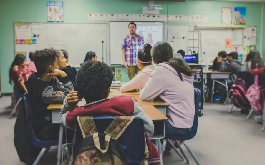 Diverse middle school students seated at desks face a male teacher presenting a lesson on a smartboard.