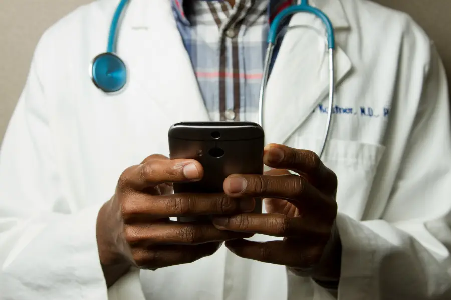 Medical professional in white lab coat and stethoscope using a smartphone; viewed from chest down.