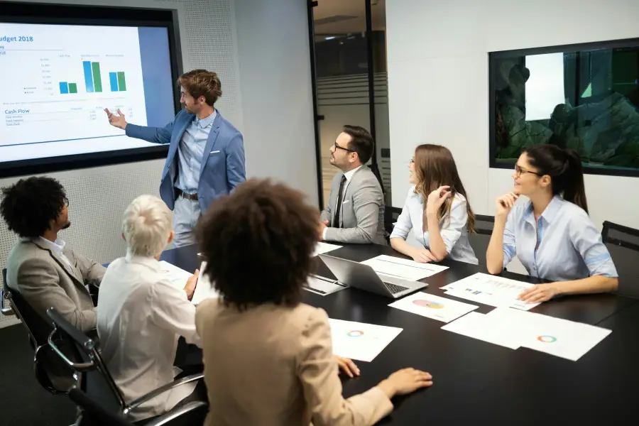 Business professional presents 2018 budget cash flow charts to a diverse team of six colleagues in a modern conference room.