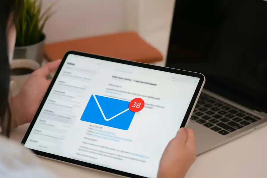 Professional reviewing an email inbox with 38 unread messages on a tablet; with a laptop in the background.