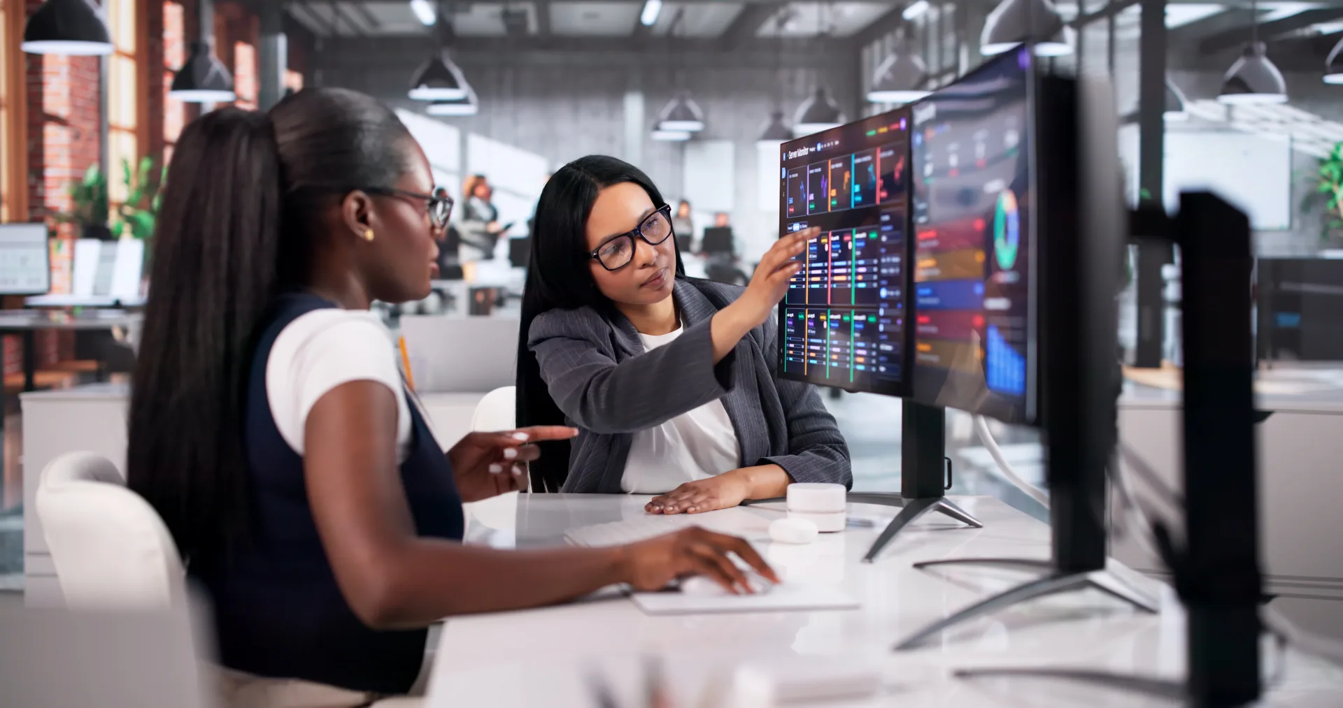 Two professional women reviewing colorful data dashboards on large monitors in a modern office environment.