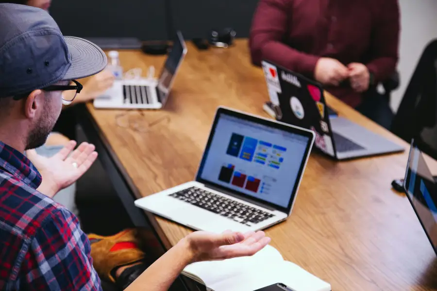 IT professionals collaborating around a wooden table with multiple laptops displaying dashboards during a team strategy me...
