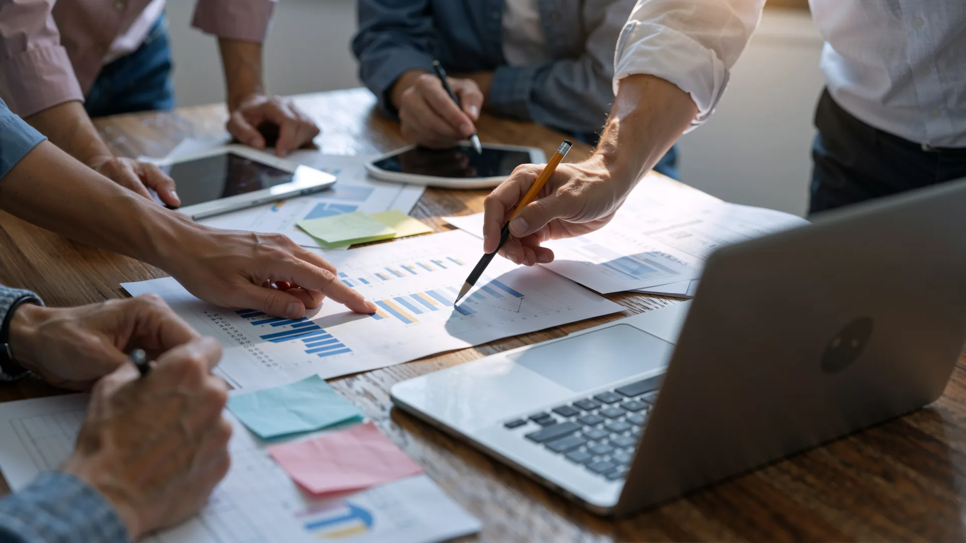 Business professionals reviewing charts; graphs; and data reports on a wooden conference table with laptops and tablets.