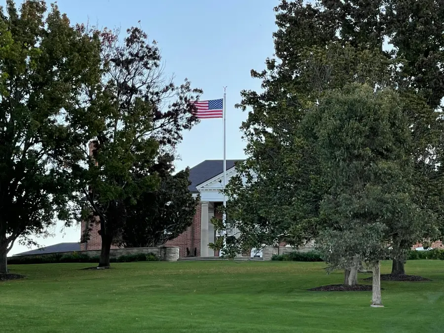 American flag flying above a brick government building with white columns; surrounded by mature trees and manicured lawn.
