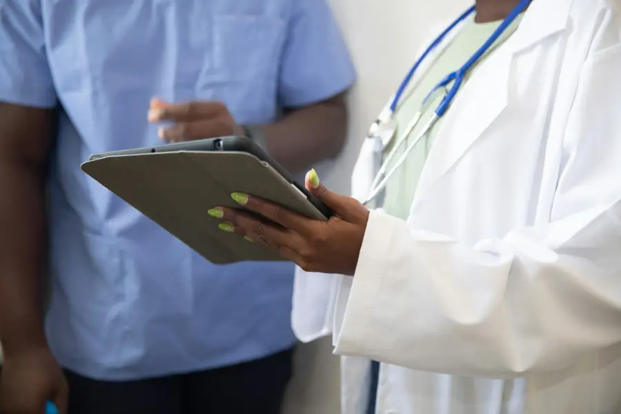 Doctor in white lab coat holding a tablet consults with a colleague in blue scrubs in a clinical setting.
