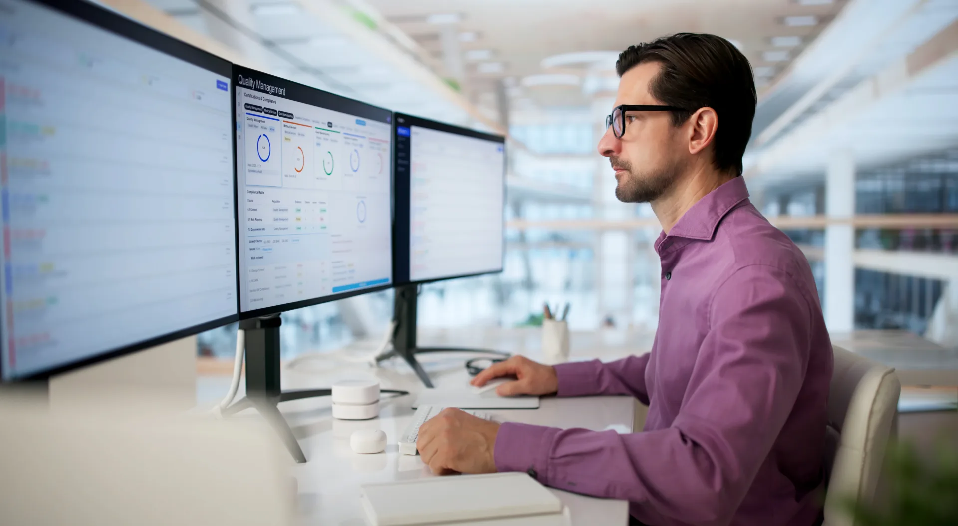 IT professional in purple shirt reviews Quality Management compliance dashboard across multiple monitors in modern office.