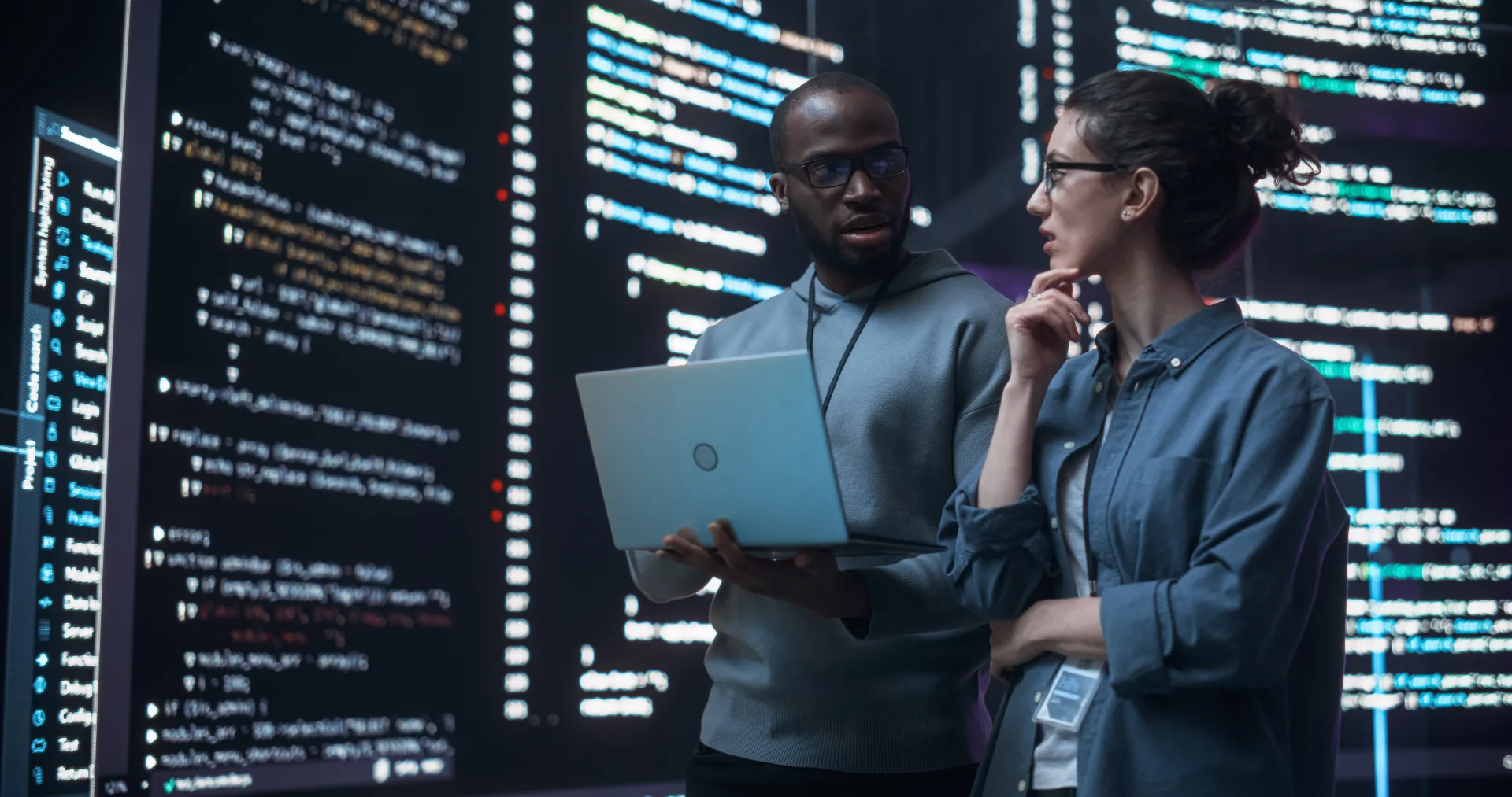 Two IT professionals reviewing source code on a laptop in front of large screens displaying software code in a dark server...