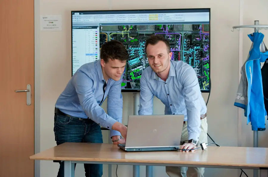 Two professionals in button-down shirts review GIS mapping software on a laptop with a large network map displayed on scre...