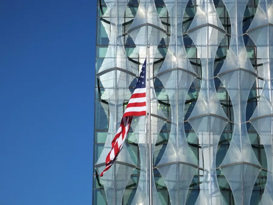 American flag flying in front of the geometric glass facade of the US Embassy in London under a clear blue sky.