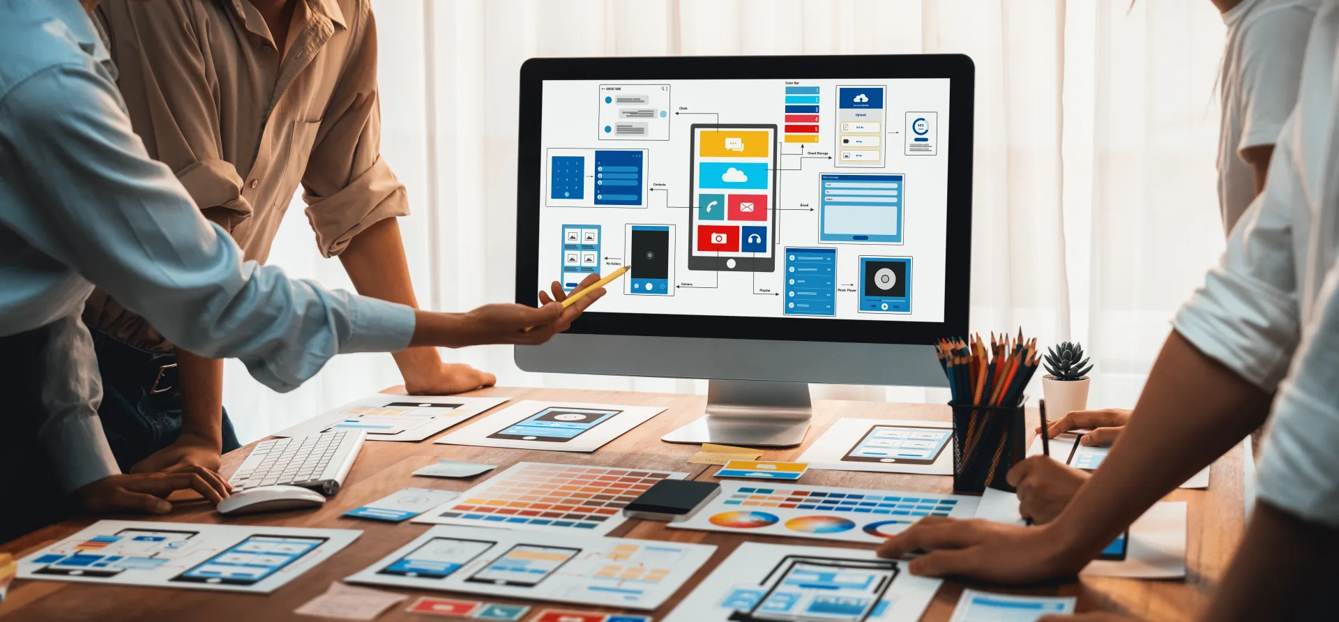 Designers gather around a monitor showing app wireframes, with printed mockups and color swatches spread across the desk.