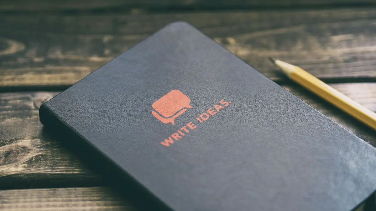 A closed notebook lettered "Write ideas" lying on a wooden desk beside a pencil.