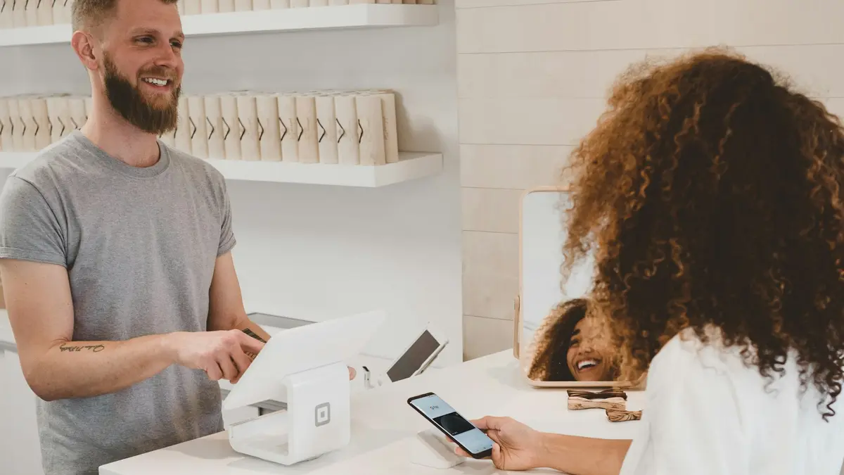 A shopkeeper stands at a small retail counter as a customer pays using a phone.