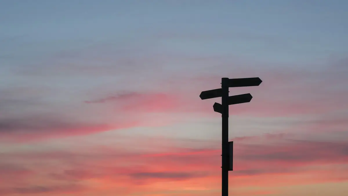 A fingerpost with arms pointing in several directions, silhouetted against a pink sunset sky.