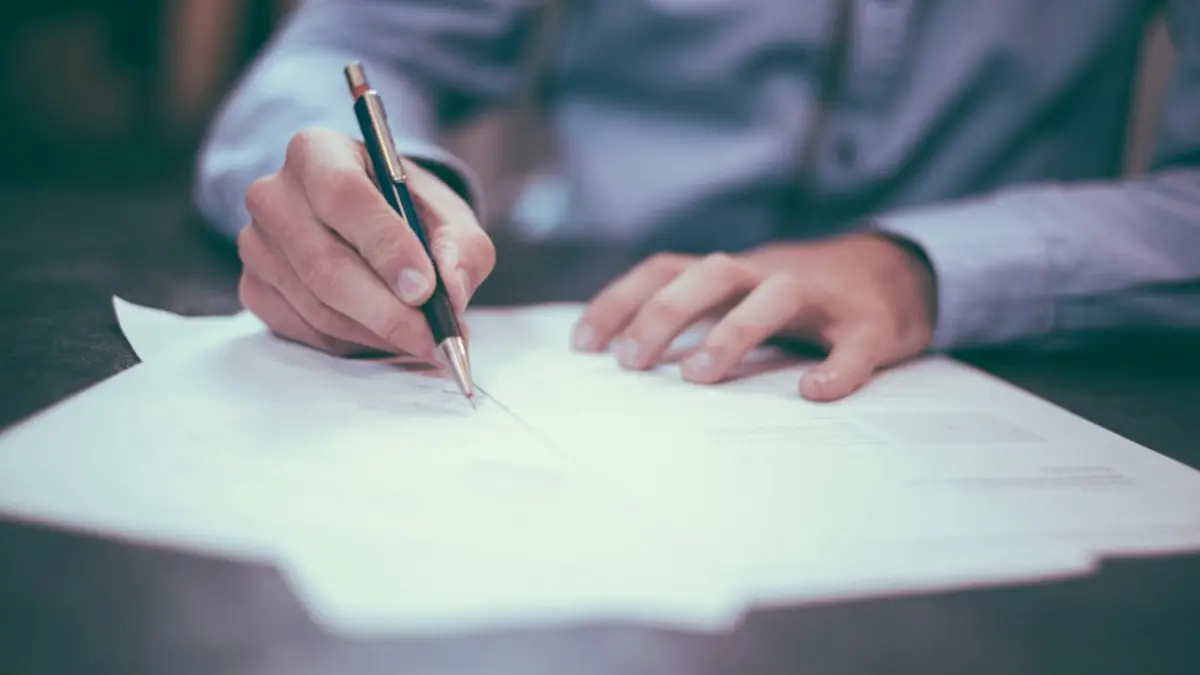 A person leaning over a desk, pen in hand, signing a sheet of paper.
