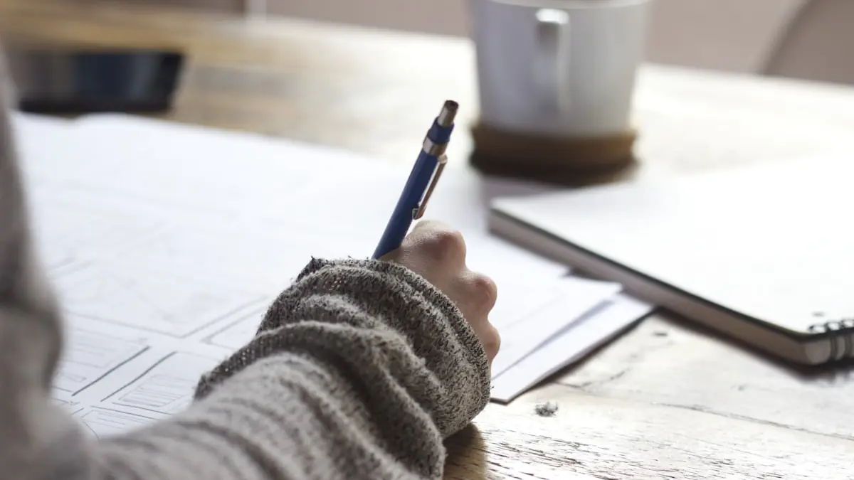 A person writing on a printed document at a wooden desk beside a cup of coffee.