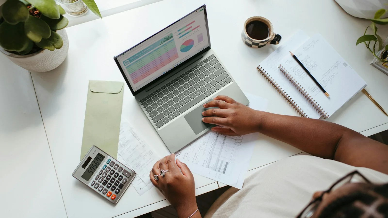 An overhead view of a desk with a laptop showing a budget spreadsheet, a calculator, and a notebook.