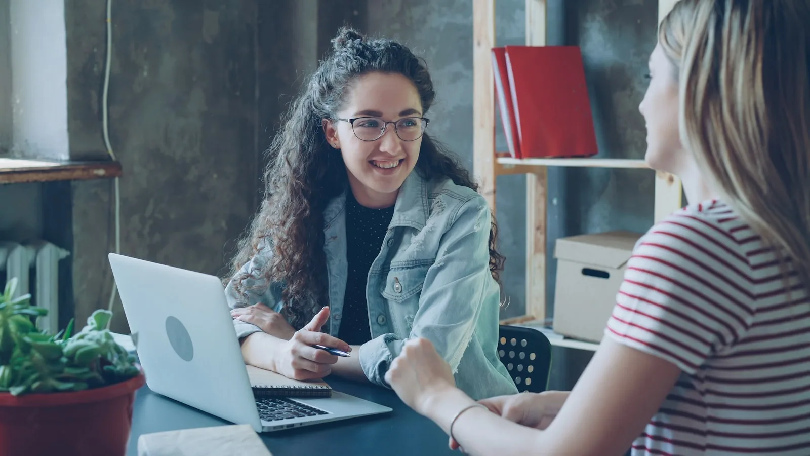 Two colleagues talking over an open laptop at a table in a small workspace.