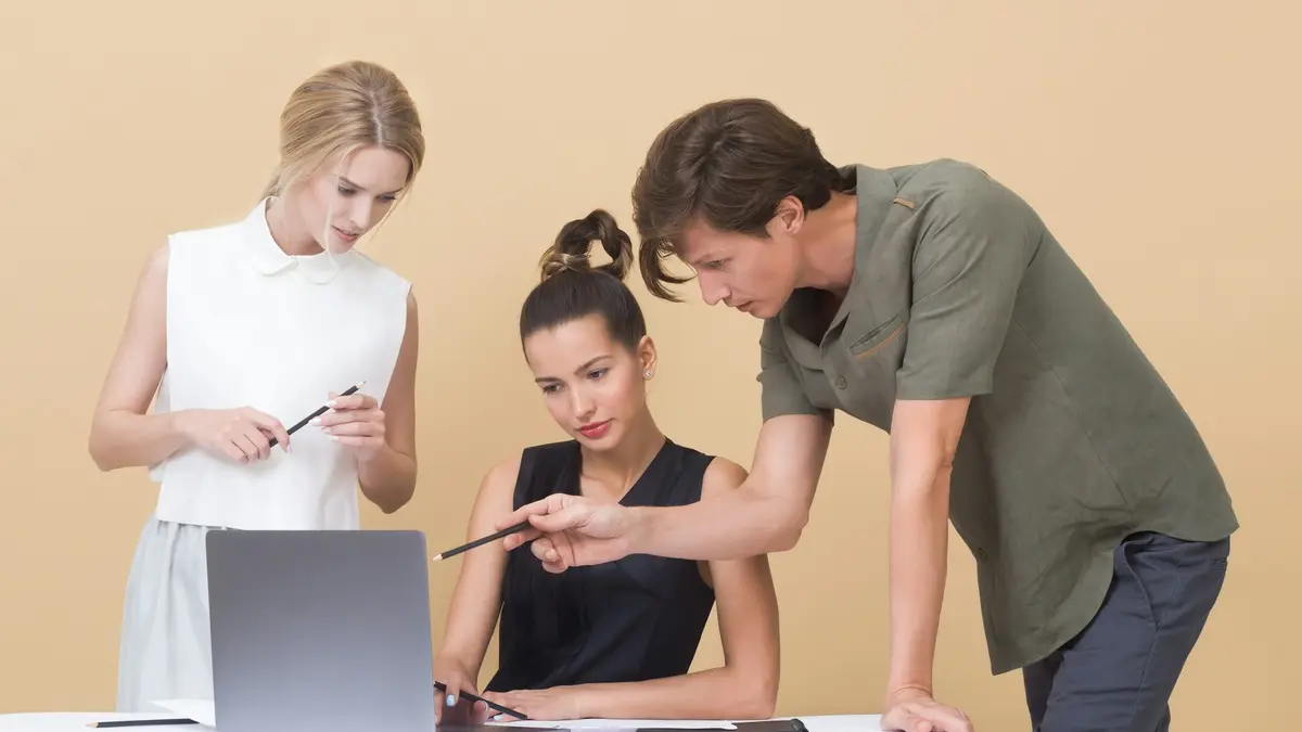 Three colleagues gathered around a laptop, one pointing at the screen while explaining.