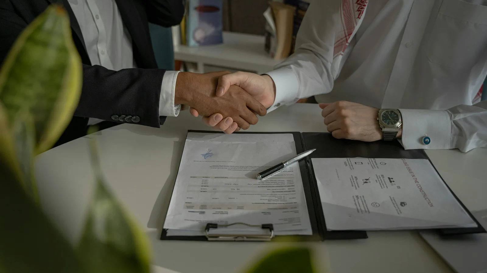 Two people shaking hands over a signed document folder and pen on a desk.