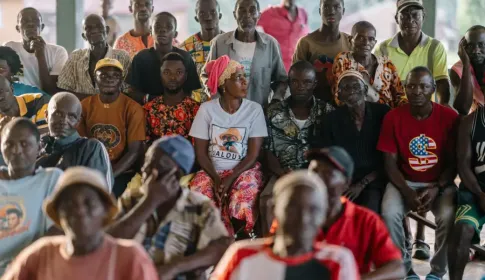 A large group of men and women of various ages seated and standing together at an outdoor community meeting.