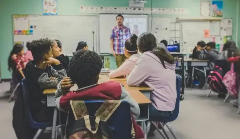 Diverse middle school students seated at desks face a male teacher presenting a lesson on a smartboard.