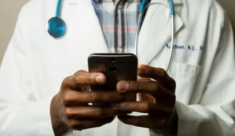 Medical professional in white lab coat and stethoscope using a smartphone; viewed from chest down.