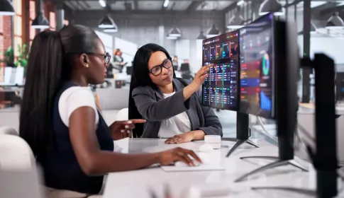 Two professional women reviewing colorful data dashboards on large monitors in a modern office environment.