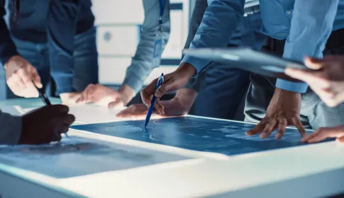 Team of professionals collaborating over a glowing digital display table; reviewing technical diagrams in an office setting.