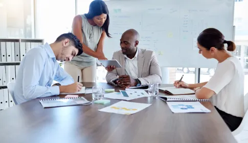 Four professionals collaborate at a conference table with charts and a tablet, a business analysis whiteboard behind them.