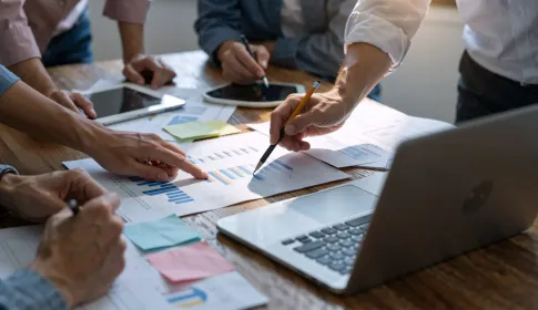 Business professionals reviewing charts; graphs; and data reports on a wooden conference table with laptops and tablets.