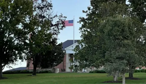 American flag flying above a brick government building with white columns; surrounded by mature trees and manicured lawn.