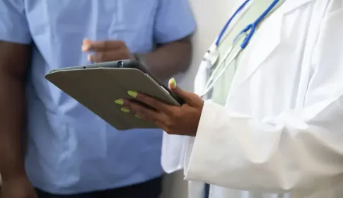 Doctor in white lab coat holding a tablet consults with a colleague in blue scrubs in a clinical setting.