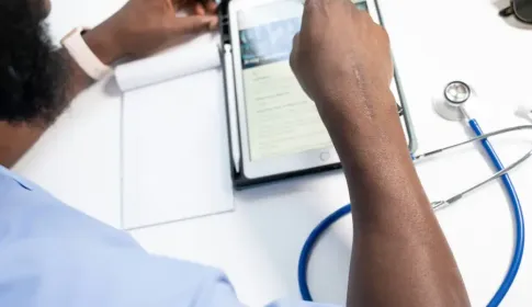 Medical professional in scrubs reviewing patient records on a tablet beside a stethoscope and notepad at a desk.