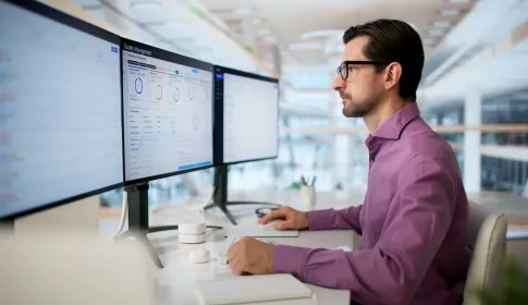 IT professional in purple shirt reviews Quality Management compliance dashboard across multiple monitors in modern office.