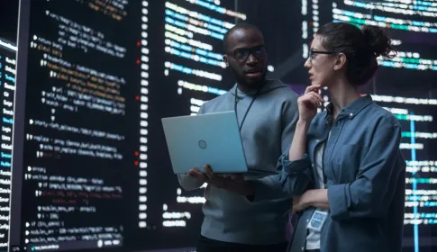 Two IT professionals review source code on a laptop in front of large screens displaying code in a dark room.