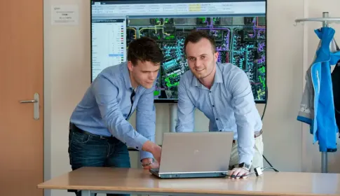 Two professionals in button-down shirts review GIS mapping software on a laptop, with a large network map on screen.