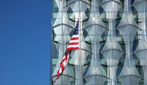 American flag flying in front of the geometric glass facade of the US Embassy in London under a clear blue sky.
