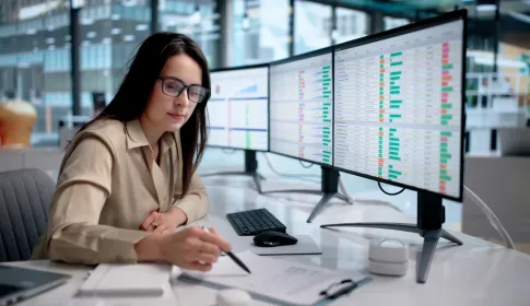 Woman in glasses reviews data dashboards on dual monitors at an enterprise office desk; taking notes with a pen.