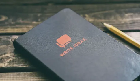 A closed notebook lettered "Write ideas" lying on a wooden desk beside a pencil.