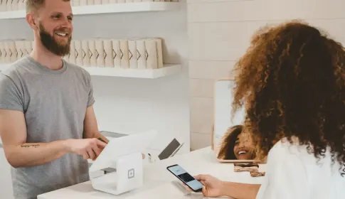 A shopkeeper stands at a small retail counter as a customer pays using a phone.