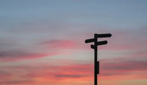 A fingerpost with arms pointing in several directions, silhouetted against a pink sunset sky.