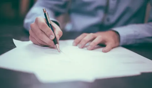 A person leaning over a desk, pen in hand, signing a sheet of paper.