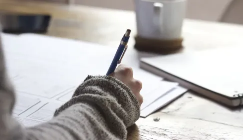 A person writing on a printed document at a wooden desk beside a cup of coffee.