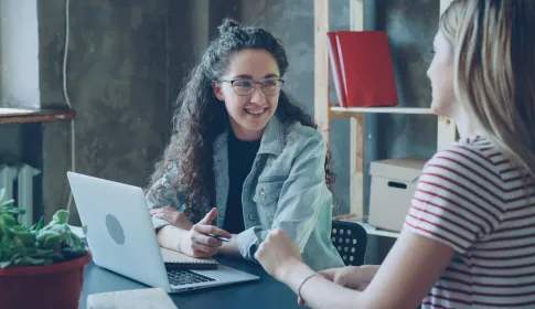 Two colleagues talking over an open laptop at a table in a small workspace.