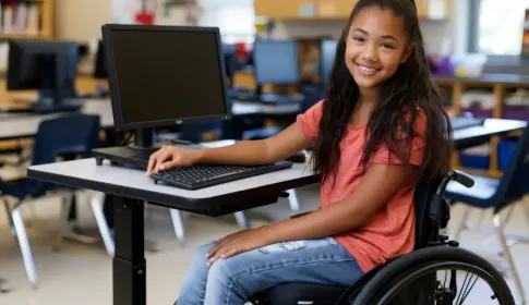 Smiling middle school student in a wheelchair uses a desktop computer at an accessible desk in a K-12 classroom.