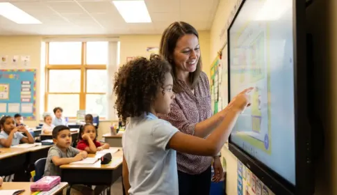 A teacher guides a young student using a touchscreen interactive whiteboard while classmates watch from their desks.