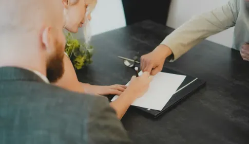 A person signing a document on a folder while two colleagues look on across a meeting table.