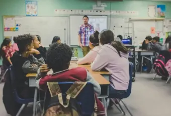 Diverse middle school students seated at desks face a male teacher presenting a lesson on a smartboard.